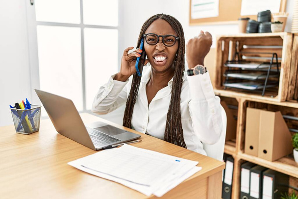 Black woman with braids working at the office speaking on the phone angry and mad raising fist frustrated and furious while shouting with anger. rage and aggressive concept.  - 写真・画像