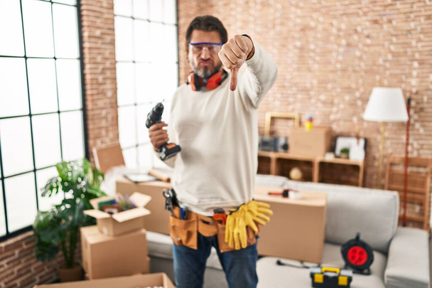 Handsome middle age man holding screwdriver at new home looking unhappy and angry showing rejection and negative with thumbs down gesture. bad expression.  - Foto, afbeelding