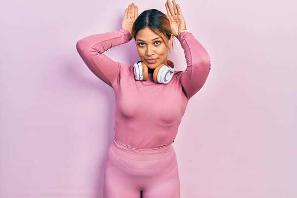 Beautiful hispanic woman wearing gym clothes and using headphones doing bunny ears gesture with hands palms looking cynical and skeptical. easter rabbit concept.  - Photo, Image