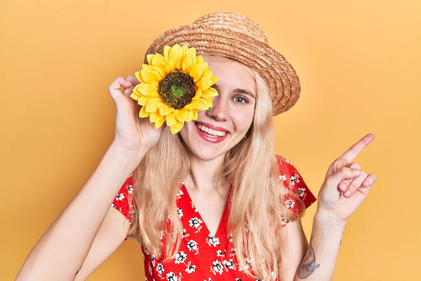 Όμορφη καυκάσια γυναίκα με ξανθά μαλλιά που κρατά κίτρινα ηλιοτρόπια πάνω από το μάτι χαμογελώντας ευτυχισμένη δείχνοντας με το χέρι και το δάχτυλο στο πλάι  - Φωτογραφία, εικόνα