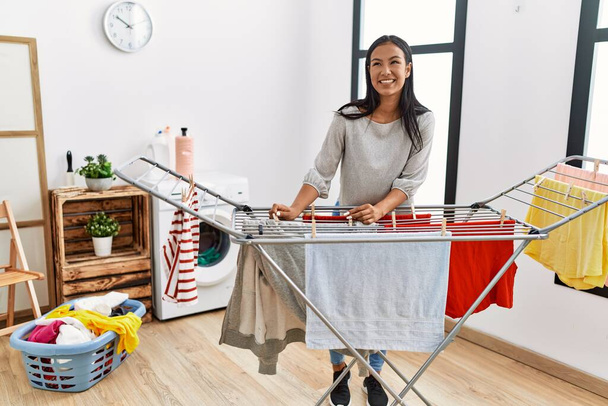 Mujer latina joven sonriendo confiada colgando ropa en la lavandería - Foto, Imagen