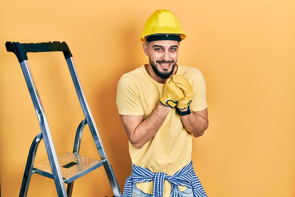 Όμορφος άντρας με γενειάδα από κατασκευαστικές σκάλες φορώντας καπέλο γελώντας νευρικός και ενθουσιασμένος με τα χέρια στο πηγούνι κοιτάζοντας στο πλάι  - Φωτογραφία, εικόνα