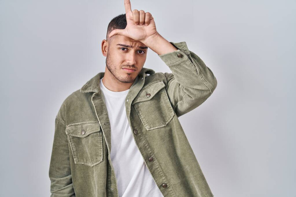 Young hispanic man standing over isolated background making fun of people with fingers on forehead doing loser gesture mocking and insulting.  - Photo, Image