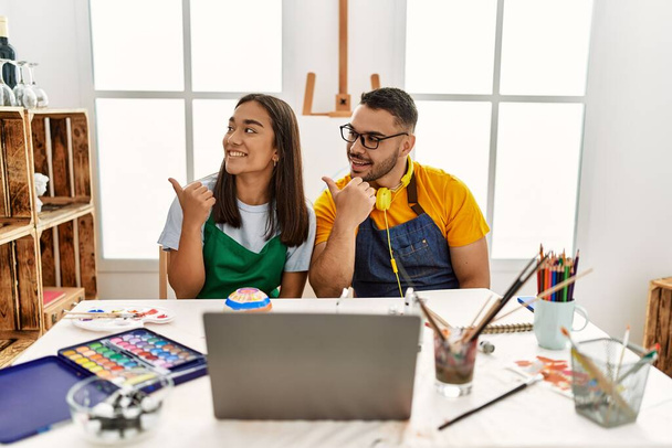 Young hispanic couple using laptop sitting on the table at art studio smiling with happy face looking and pointing to the side with thumb up.  - Foto, Imagen