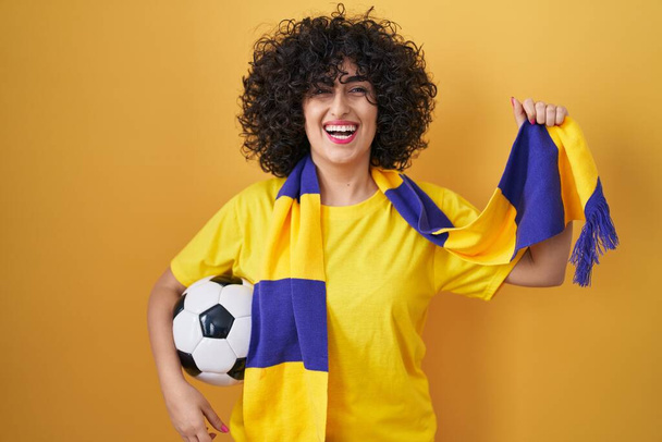 Νεαρή μελαχρινή γυναίκα με σγουρά μαλλιά χούλιγκαν ποδόσφαιρο κρατώντας μπάλα χαμογελώντας και γελώντας δυνατά γιατί αστείο τρελό αστείο.  - Φωτογραφία, εικόνα