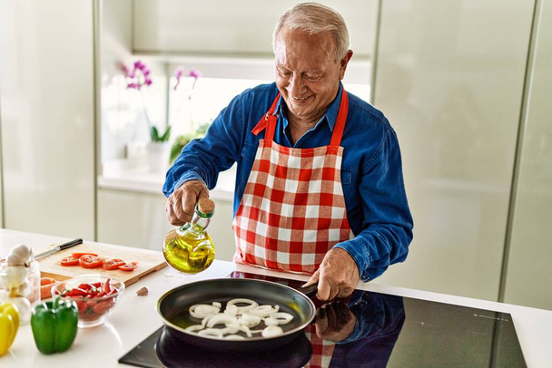 Senior άνθρωπος χαμογελά αυτοπεποίθηση ρίχνει λάδι στο τηγάνι στην κουζίνα - Φωτογραφία, εικόνα