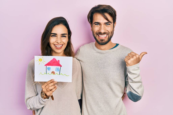 Young hispanic couple holding house draw pointing thumb up to the side smiling happy with open mouth  - Foto, afbeelding