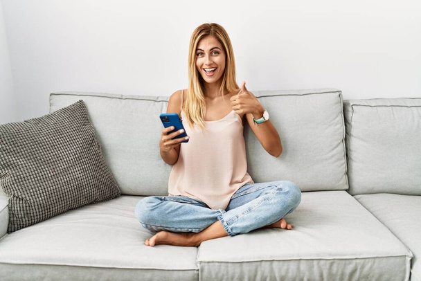 Szőke gyönyörű fiatal nő ül a kanapén otthon okostelefonnal csinál boldog hüvelykujj fel gesztus kézzel. jóváhagyása kifejezés nézi a kamera mutatja siker.  - Fotó, kép