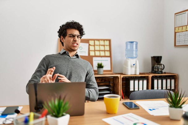 Νεαρός ισπανόφωνος άνδρας που φοράει επιχειρηματικό στυλ κάθεται στο γραφείο αηδιασμένος έκφραση, δυσαρεστημένος και φοβισμένος να κάνει το πρόσωπο αηδίας επειδή η αντίδραση αποστροφής. με τα χέρια σηκωμένα  - Φωτογραφία, εικόνα
