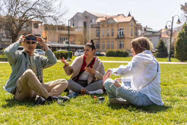 Venkovní záběr šťastných mladých přátel sedících spolu na trávníku. Muži a ženy si užívají den na louce - Fotografie, Obrázek