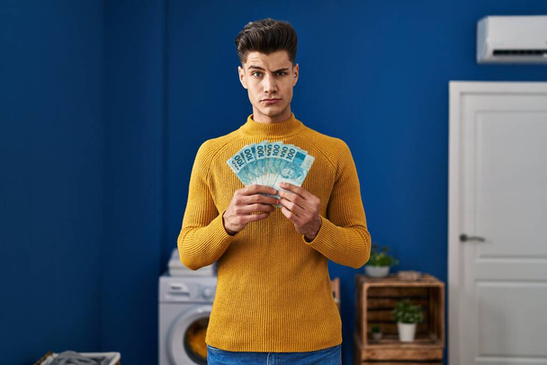 Young hispanic man at laundry room holding brazilian reals skeptic and nervous, frowning upset because of problem. negative person.  - Fotografie, Obrázek
