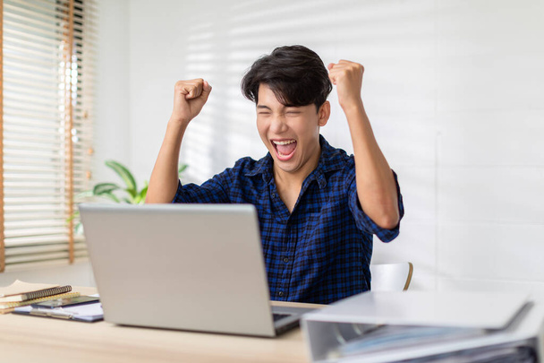 アジア人男性は、コンピュータのラップトップ画面上で笑顔と幸せな気持ちを表現しています。若い男性は良いニュースを得たと彼の陽気な顔を表示します。 - 写真・画像