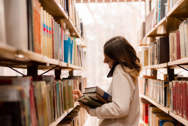 Meisje kiest een boek uit de boekenplanken in de bibliotheek terwijl ze telefoneert - Foto, afbeelding