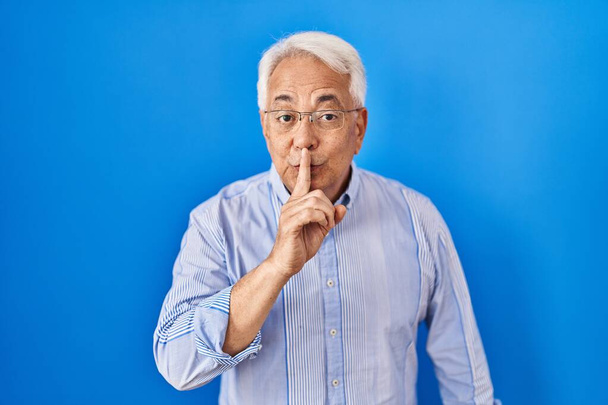 Spaanse senior man draagt een bril en vraagt om stil te zijn met de vinger op de lippen. stilte en geheim concept.  - Foto, afbeelding