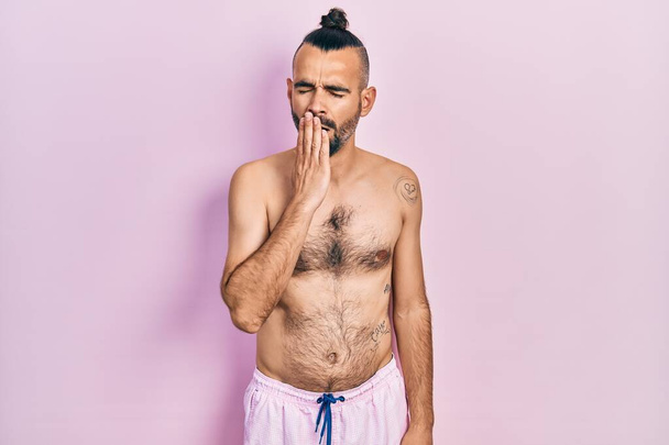 Junge hispanische Mann ohne Hemd und Badeanzug gelangweilt gähnende müde Mund mit der Hand bedecken. Ruhelos und schläfrig.  - Foto, Bild