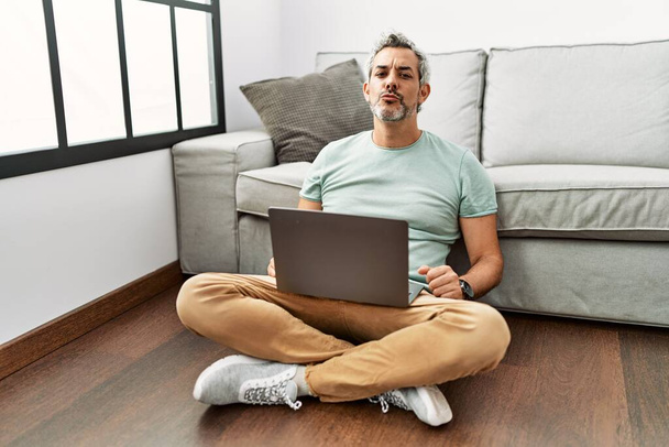 Hombre hispano de mediana edad usando portátil sentado en el suelo en la sala de estar mirando a la cámara soplando un beso en el aire siendo encantador y sexy. expresión de amor.  - Foto, imagen