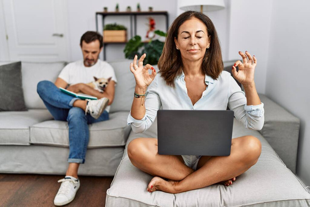 Hispanic middle age couple at home, woman using laptop relax and smiling with eyes closed doing meditation gesture with fingers. yoga concept.  - Φωτογραφία, εικόνα