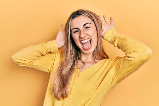 Красива іспаномовна жінка в повсякденному жовтому светрі посміхається весело грати в пошуках боу з руками, що показують обличчя. здивований і вийшов
  - Фото, зображення