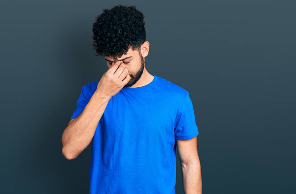 Młody arabski mężczyzna z brodą w luźnej niebieskiej koszuli zmęczony pocieraniem nosa i oczu uczucie zmęczenia i bólu głowy. koncepcja stresu i frustracji.  - Zdjęcie, obraz
