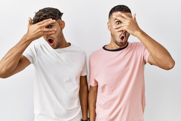 Mladý homosexuální pár stojící nad izolovaným pozadím vykukující v šoku a zakrývající obličej a oči rukou, dívající se skrz prsty s rozpačitým výrazem.  - Fotografie, Obrázek