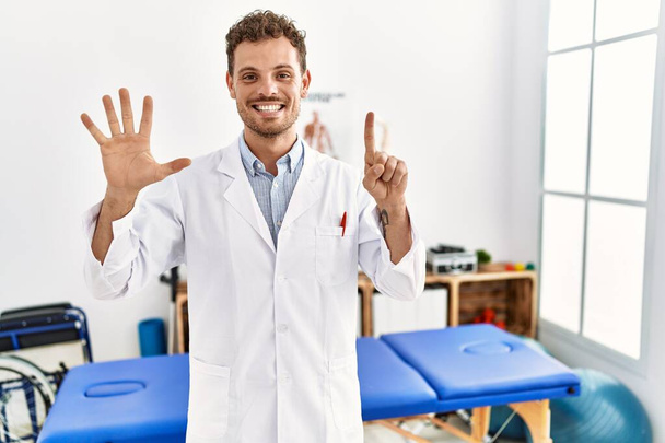 Handsome young man working at pain recovery clinic showing and pointing up with fingers number six while smiling confident and happy.  - Photo, image