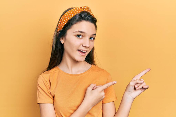 Νεαρή καστανή κοπέλα που φοράει πορτοκαλί μπλουζάκι χαμογελώντας και κοιτάζοντας την κάμερα που δείχνει με τα δύο χέρια και τα δάχτυλα στο πλάι.  - Φωτογραφία, εικόνα