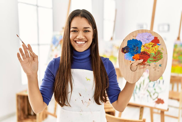 Jonge latijnse vrouw glimlachend zelfverzekerd met penseel en palet in kunststudio - Foto, afbeelding