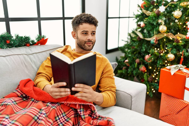 Young arab man reading book lying on the sofa by christmas tree at home. - Zdjęcie, obraz