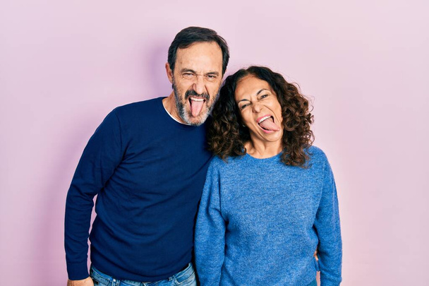 Middelbare leeftijd paar Latijns-Amerikaanse vrouw en man knuffelen en staan samen steken tong uit gelukkig met grappige uitdrukking. emotie concept.  - Foto, afbeelding