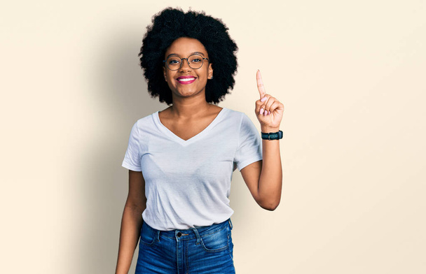 Jeune femme afro-américaine portant t-shirt blanc décontracté montrant et pointant vers le haut avec le doigt numéro un tout en souriant confiant et heureux.  - Photo, image