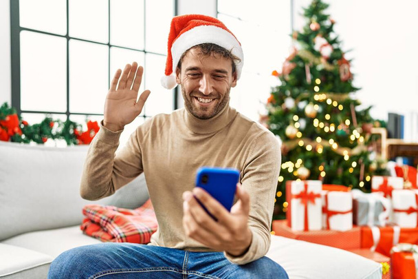 Giovane uomo ispanico avendo videochiamata seduto vicino all'albero di Natale a casa - Foto, immagini