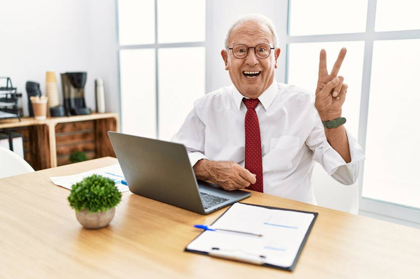 Senior man working at the office using computer laptop smiling with happy face winking at the camera doing victory sign with fingers. number two.  - Foto, Imagem