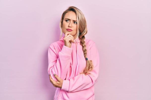 Schöne junge blonde Frau in rosa Sweatshirt, die konzentriert über Zweifel nachdenkt, mit dem Finger am Kinn und fragend nach oben schaut  - Foto, Bild