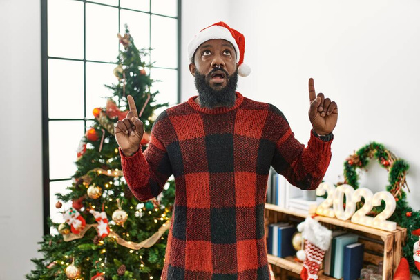 Αφροαμερικάνος που φορούσε καπέλο Σάντα Κλάους και στεκόταν δίπλα στο χριστουγεννιάτικο δέντρο έκπληκτος που κοίταζε ψηλά και έδειχνε με τα δάχτυλα και σήκωνε τα χέρια..  - Φωτογραφία, εικόνα