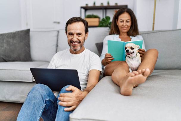 Middle age man and woman couple using laptop and reading book sitting on sofa with chihuahua at home - Photo, Image