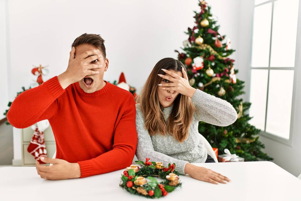 Young hispanic couple sitting at the table on christmas peeking in shock covering face and eyes with hand, looking through fingers with embarrassed expression.  - Photo, Image