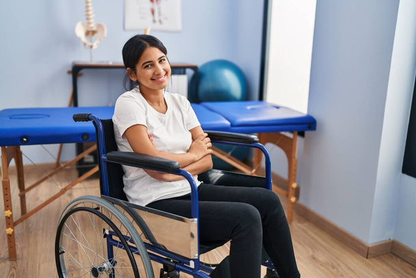 Νεαρή Ισπανίδα που κάθεται σε αναπηρική καρέκλα στην κλινική φυσιοθεραπείας, χαρούμενο πρόσωπο χαμογελώντας με σταυρωμένα τα χέρια κοιτάζοντας την κάμερα. θετικό πρόσωπο.  - Φωτογραφία, εικόνα