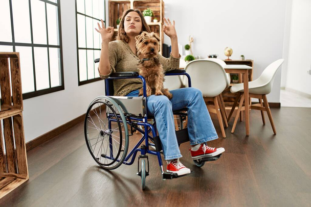 Młoda Latynoska dziewczyna siedząca na wózku inwalidzkim w domu uśmiechnięta w miłości robiąca ręcznie kształt symbolu serca. romantyczna koncepcja.  - Zdjęcie, obraz