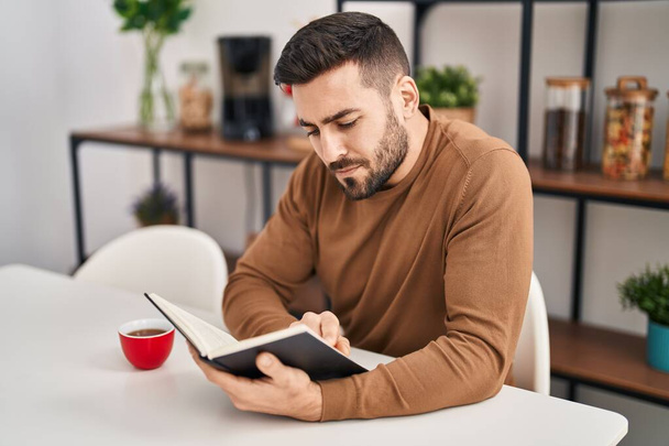 Young hispanic man reading book and drinking coffee sitting on table at home - Foto, Imagem