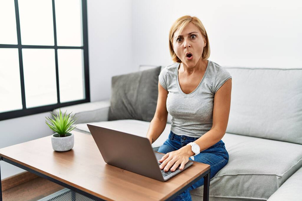 Middle age blonde woman using laptop at home in shock face, looking skeptical and sarcastic, surprised with open mouth  - Photo, Image
