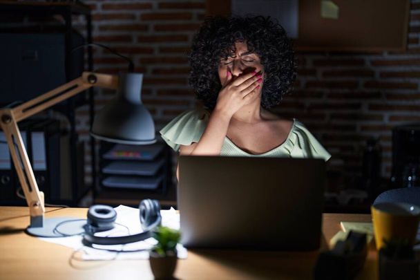 Fiatal barna nő göndör hajjal dolgozik az irodában éjszaka unatkozik ásít fáradt eltakarja száját kézzel. nyugtalanság és álmosság.  - Fotó, kép
