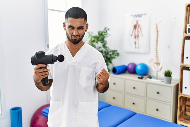 Young indian physiotherapist holding therapy massage gun at wellness center very happy and excited doing winner gesture with arms raised, smiling and screaming for success. celebration concept.  - Foto, imagen