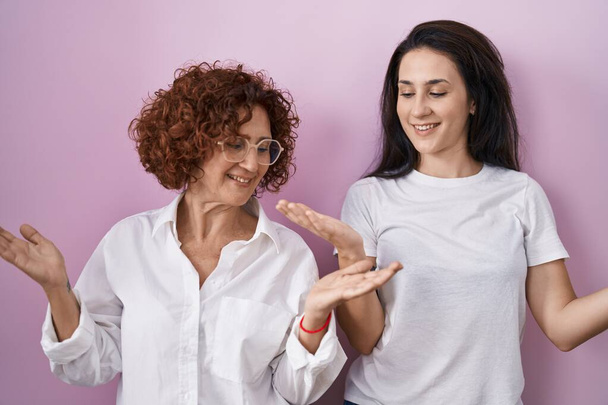 Латиноамериканская мать и дочь одеты в повседневную белую футболку на розовом фоне, улыбаясь, показывая обе руки открытыми ладонями, представляя и рекламу сравнение и баланс  - Фото, изображение