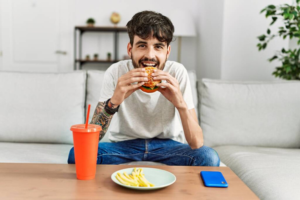 Joven hispano comiendo hamburguesa clásica y bebiendo refresco sentado en el sofá en casa. - Foto, imagen