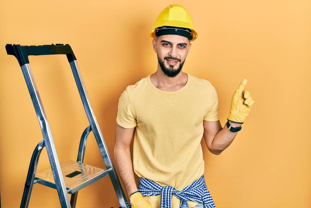 Handsome man with beard by construction stairs wearing hardhat with a big smile on face, pointing with hand finger to the side looking at the camera.  - Foto, imagen