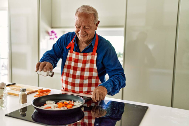 Idősebb férfi mosolyog magabiztos öntő só serpenyőben a konyhában - Fotó, kép