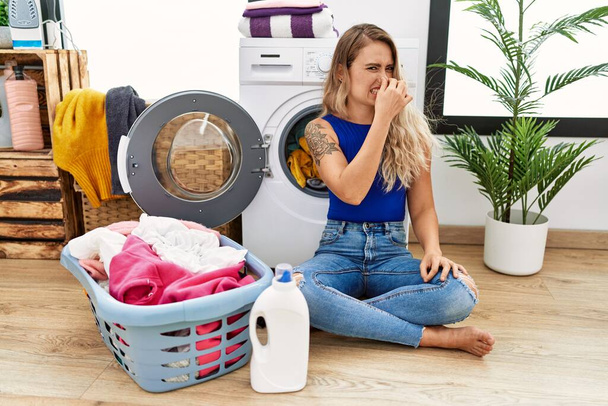 Young beautiful woman doing laundry sitting by wicker basket smelling something stinky and disgusting, intolerable smell, holding breath with fingers on nose. bad smell  - Photo, Image
