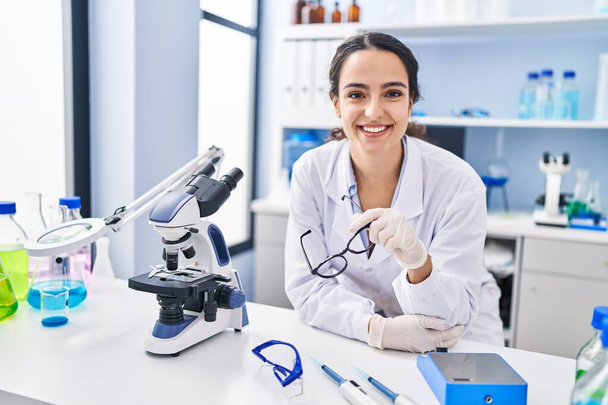 Молодая латиноамериканка в научной форме, уверенно улыбающаяся лаборатории - Фото, изображение