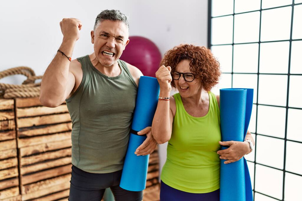 Middle age couple holding yoga mat angry and mad raising fist frustrated and furious while shouting with anger. rage and aggressive concept.  - 写真・画像
