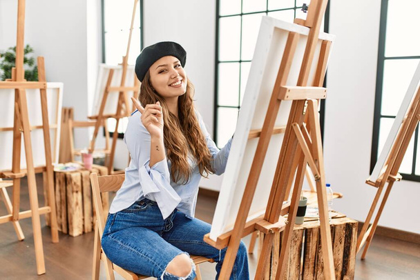 Junge hispanische Künstlerin malt auf Leinwand im Kunstatelier mit einem breiten Lächeln im Gesicht und zeigt mit dem Finger zur Seite in die Kamera.  - Foto, Bild